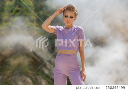Woman with short blonde hair posing in a purple outfit against a steamy geothermal landscape Woman with short blonde hair posing in a purple outfit against a steamy geothermal landscape 138846499