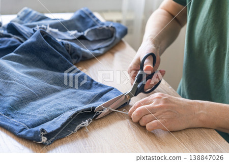 Hands cutting thread from old jeans for reuse, promoting sustainable fashion and circular economy principles. Person upcycling denim jeans with scissors, repairing clothing 138847026