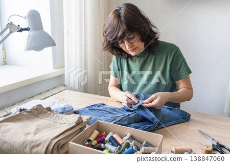 Woman mending blue denim pants by hand, focusing on sustainable clothing repair and textile handicraft at home. An adult woman repairing clothes. Reusing things Woman mending blue denim pants by hand, focusing on sustainable clothing repair and textile handicraft at home. An adult woman repairing clothes. Reusing things 138847060