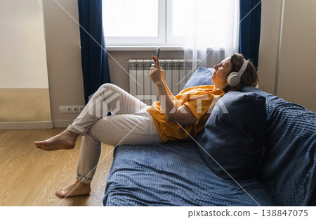 Woman lying on couch wearing white wireless headphones, enjoying audio from smartphone, surfing the internet in a cozy living room Woman lying on couch wearing white wireless headphones, enjoying audio from smartphone, surfing the internet in a cozy living room 138847075
