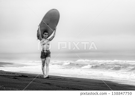 Strong woman with dramatic makeup holds surfboard on her head black sand beach 138847750