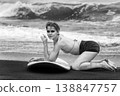 Monochrome fashion portrait of young woman with surfboard kneeling on dark sand beach 138847757