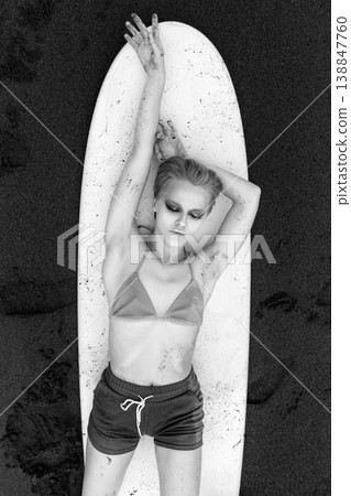 Monochrome top view of woman resting on surfboard on black sand beach, lying on back Monochrome top view of woman resting on surfboard on black sand beach, lying on back 138847760