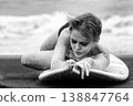 Woman with thoughtful expression lying on surfboard on sandy beach. Black and white 138847764