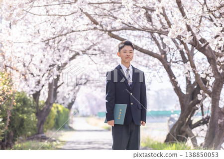 A male student stands under a cherry tree, holding his graduation certificate. 138850053