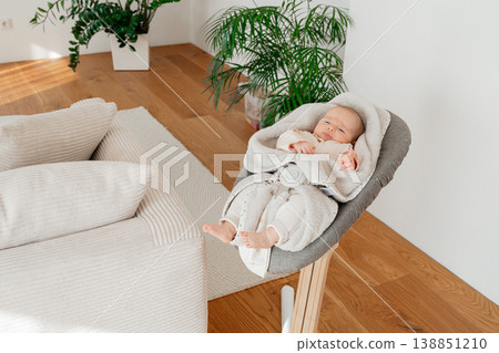 Baby relaxing in a bouncer near sofa in modern living room. Warm sunlight and cozy home environment. Family lifestyle and childcare concept. Baby relaxing in a bouncer near sofa in modern living room. Warm sunlight and cozy home environment. Family lifestyle and childcare concept. 138851210