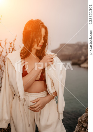 Woman coast hoodie posing on a rocky shore wearing a white tracksuit and bikini top during a golden sunset at the sea Woman coast hoodie posing on a rocky shore wearing a white tracksuit and bikini top during a golden sunset at the sea 138851501