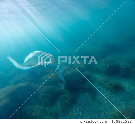 Freediver ocean sunbeams woman in long fins diving into deep blue water towards a rocky seabed under mystical rays of sunlight 138851588
