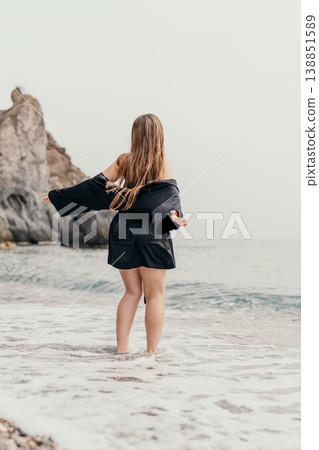 Woman beach robe standing on misty shoreline with arms outstretched enjoying peaceful ocean breeze and rocky cliffs view 138851589