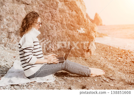 Woman laptop beach working remotely on a rocky coastline wearing a striped sweater during sunset for a digital nomad lifestyle Woman laptop beach working remotely on a rocky coastline wearing a striped sweater during sunset for a digital nomad lifestyle 138851608