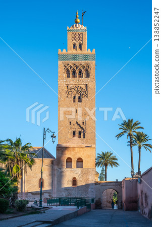 Kutubiyya Mosque is visible in Marrakesh. The mosque features a tall minaret surrounded by palm trees. People walk nearby, enjoying the sunny day in a lively area of the city. Kutubiyya Mosque is visible in Marrakesh. The mosque features a tall minaret surrounded by palm trees. People walk nearby, enjoying the sunny day in a lively area of the city. 138852247