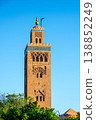 The tower of Kutubiyya Mosque stands tall in Marrakesh, Morocco. The clear blue sky forms a backdrop as the sun shines on this historical site. Visitors may be nearby, enjoying the scene. 138852249