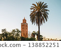 Kutubiyya Mosque is located in Marrakesh, Morocco, surrounded by palm trees. Visitors admire the scene as the sun begins to set in the sky, creating a lively atmosphere. 138852251