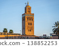 The tower of Koutoubia Mosque stands tall in Marrakesh. The sun sets behind it, casting warm light on the structure. Nearby palm trees add to the scene, creating a lively atmosphere. 138852253