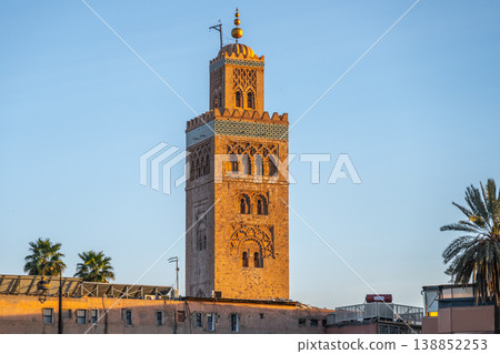 The tower of Koutoubia Mosque stands tall in Marrakesh. The sun sets behind it, casting warm light on the structure. Nearby palm trees add to the scene, creating a lively atmosphere. 138852253
