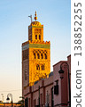 The Kutubiyya Mosque tower is visible against the evening sky in Marrakesh. The sunlight casts warm tones on the stone as people walk nearby. The location is popular among visitors. 138852255