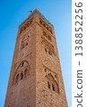 People gather around Kutubiyya Mosque in Marrakesh, Morocco. The sun shines brightly on the tall minaret. Visitors admire the historical architecture and take pictures. 138852256
