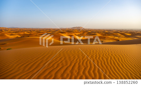 In the Sahara Desert, the vast sand dunes of Erg Chebbi stretch under a clear blue sky. The sun shines on the golden sands, highlighting the natural patterns formed by the wind. 138852260