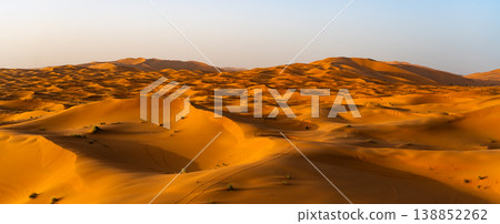 Visitors explore the vast sand dunes of Erg Chebbi in Merzouga, Sahara Desert, Morocco. The sun sets, casting golden light across the undulating landscape of fine sand. 138852262