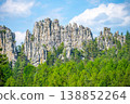 Tall rock formations stand against a blue sky filled with clouds. Green trees surround the base of the rocks. It is daytime, and the sun lights up the scene. 138852264
