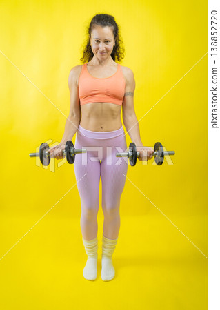 Woman exercising biceps with dumbbells on yellow background Woman exercising biceps with dumbbells on yellow background 138852720