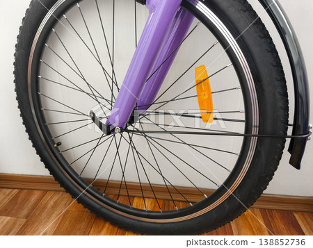 Close-up of a bicycle front wheel with black tire, metal spokes, and orange reflector on a purple frame, indoors on a wooden floor, highlighting safety, mobility, and urban lifestyle. 138852736