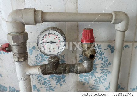 Close-up of a household plumbing system with pressure gauge, safety valve, and pipes mounted on a tiled wall, showing water pressure control equipment in a domestic setting Close-up of a household plumbing system with pressure gauge, safety valve, and pipes mounted on a tiled wall, showing water pressure control equipment in a domestic setting 138852742