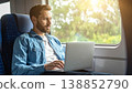 Young man working on laptop during train travel at sunset. Professional digital nomad lifestyle with golden sun flare, remote working, and commuter journey in modern railway carriage interior 138852790
