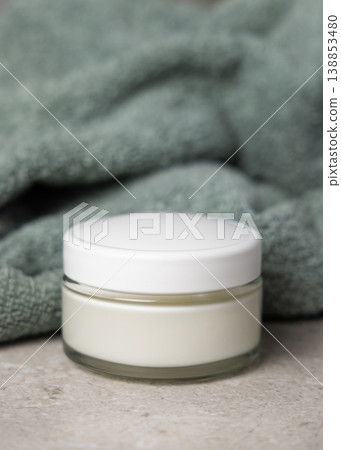 Closed cosmetic cream jar with white lid on grey stone near green leaves and bath towel mockup 138853480
