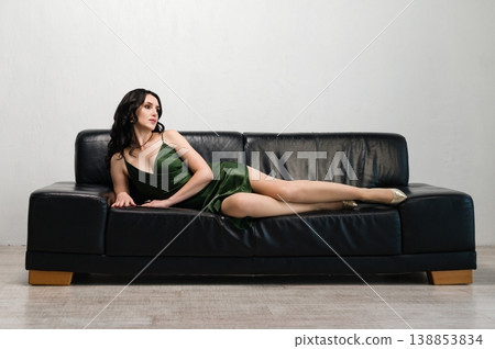 Elegant woman in a green dress relaxing on a black leather sofa. Elegant woman in a green dress relaxing on a black leather sofa. 138853834