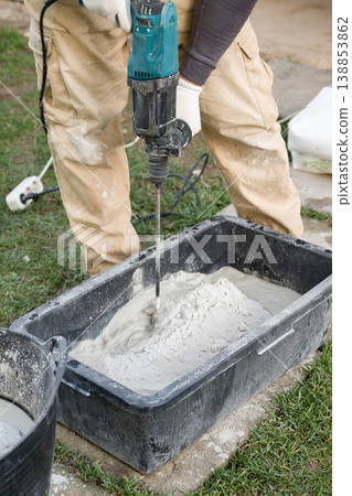 Worker mixes cement self-leveling floor in a black container using an electric drill at a construction site during daylight hours Worker mixes cement self-leveling floor in a black container using an electric drill at a construction site during daylight hours 138853862