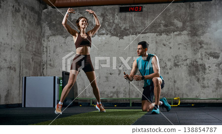 Woman performing jumping exercise while man tracks time during gym workout Woman performing jumping exercise while man tracks time during gym workout 138854787