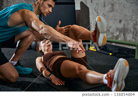 Woman performing leg raise exercise on mat with male coach guiding technique in gym. 138854804