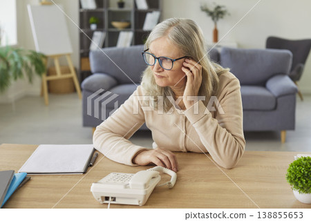 Worried sad senior woman sitting at the desk at home waiting for important phone call from someone. Worried sad senior woman sitting at the desk at home waiting for important phone call from someone. 138855653