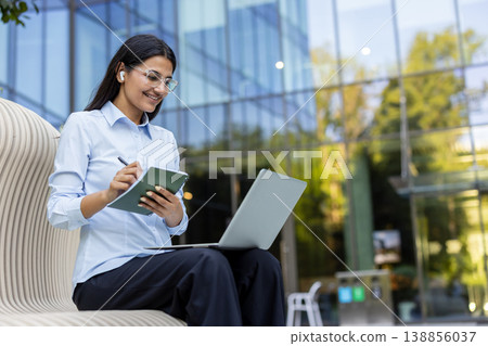 Young indian businesswoman sitting on a modern bench outdoors, happily writing notes in a planner while using a laptop and wearing wireless earphones in a remote work setting Young indian businesswoman sitting on a modern bench outdoors, happily writing notes in a planner while using a laptop and wearing wireless earphones in a remote work setting 138856037