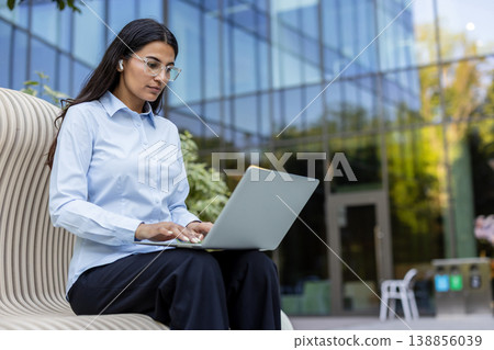 Young businesswoman typing on a laptop computer while sitting on a modern bench outside a glass office building, embracing flexible work and global connectivity 138856039