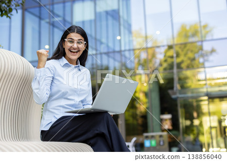 Young businesswoman smiling cheerfully, showing a fist pump gesture, and holding a laptop while sitting on a bench outside a modern glass office building, celebrating business success Young businesswoman smiling cheerfully, showing a fist pump gesture, and holding a laptop while sitting on a bench outside a modern glass office building, celebrating business success 138856040