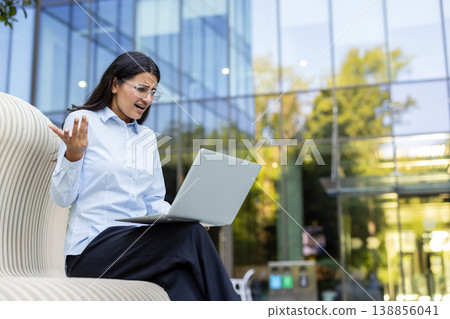 Young business woman experiencing stress and frustration while working on a laptop computer outside a modern office building, encountering a technical error or failure Young business woman experiencing stress and frustration while working on a laptop computer outside a modern office building, encountering a technical error or failure 138856041
