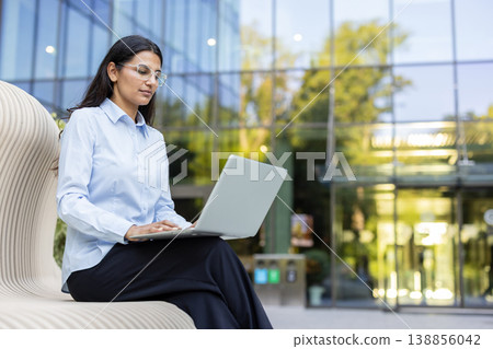 Young indian businesswoman sitting on a contemporary bench outside a corporate glass building, typing on her laptop, focusing on work and productivity in a professional urban setting 138856042