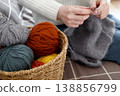 Woman in white sweater knitting with needles beside basket of colorful yarn creating cozy handmade fabric 138856799
