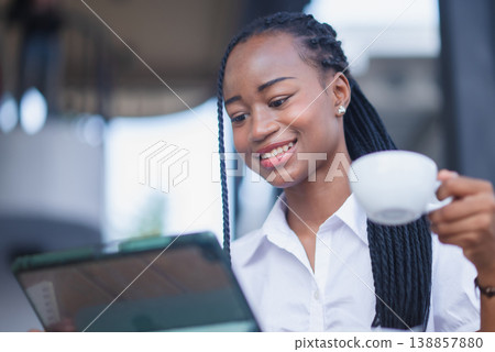 Young Black woman smiling while holding coffee and using a tablet in a bright cafe. Casual digital lifestyle, online learning, mobile browsing, remote work and morning routine concept. Young Black woman smiling while holding coffee and using a tablet in a bright cafe. Casual digital lifestyle, online learning, mobile browsing, remote work and morning routine concept. 138857880