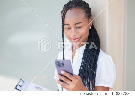 Young Black office woman checking smartphone while holding clipboard outdoors by building wall. Business communication, task planning, mobile workflow, remote work and productivity concept. Young Black office woman checking smartphone while holding clipboard outdoors by building wall. Business communication, task planning, mobile workflow, remote work and productivity concept. 138857883