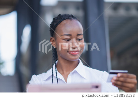 Young Black woman holding credit card and using tablet for online payment outdoors. Digital banking, ecommerce purchase, fintech transaction, cashless shopping and mobile lifestyle concept. 138858158