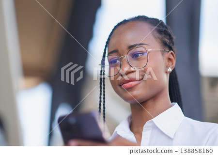 Young Black woman in glasses using smartphone outdoors with a calm smile. Mobile communication, social media browsing, online connection, digital lifestyle and modern technology concept. Young Black woman in glasses using smartphone outdoors with a calm smile. Mobile communication, social media browsing, online connection, digital lifestyle and modern technology concept. 138858160