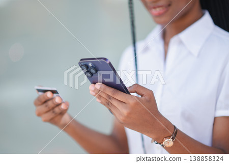 Close up of young Black woman using smartphone and bank card for online payment outdoors. Mobile banking, digital wallet, ecommerce checkout, secure transaction and cashless shopping concept. Close up of young Black woman using smartphone and bank card for online payment outdoors. Mobile banking, digital wallet, ecommerce checkout, secure transaction and cashless shopping concept. 138858324