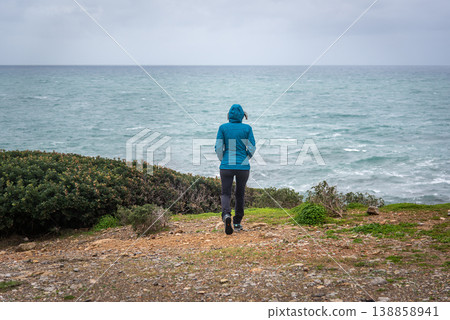 Woman in Hiking Clothes with Hood Walking Towards Stormy Coast Woman in Hiking Clothes with Hood Walking Towards Stormy Coast 138858941