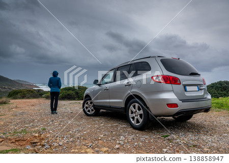 Woman in Hiking Outfit with Hood on Dirt Road near SUV by the Shore during Stormy Sea Woman in Hiking Outfit with Hood on Dirt Road near SUV by the Shore during Stormy Sea 138858947
