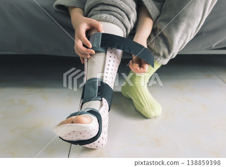 Hands adjust the strap on a patient's light pink perforated medical foot and ankle brace 138859398