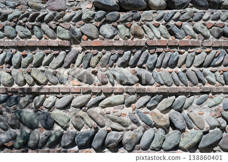 Ancient stone wall texture with rows of river pebbles laid diagonally and separated by thin brick layers. Ancient stone wall texture with rows of river pebbles laid diagonally and separated by thin brick layers. 138860401