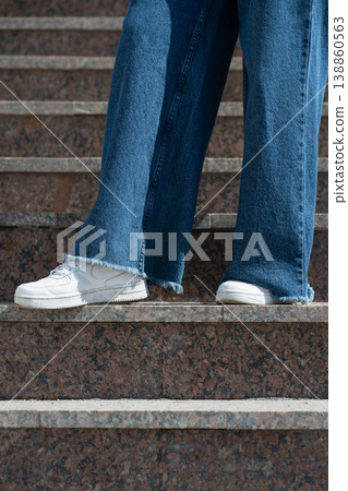 Side view of person wearing wide leg blue jeans with raw hem and white sneakers on granite stairs. 138860563